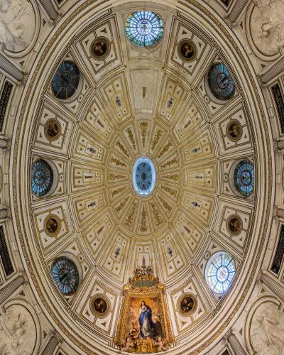 An ornate, domed interior ceiling featuring gold leaf detailing, blue stained-glass windows, and a central religious painting, viewed from below.