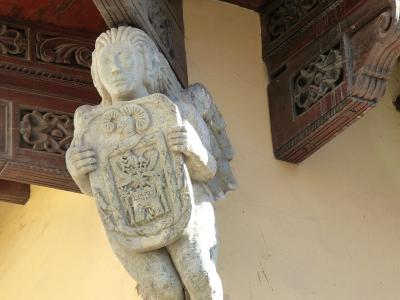A stone sculpture of a winged figure (putto or angel) holding a coat of arms, mounted under a wooden balcony with carved corbels against a yellow stucco wall.