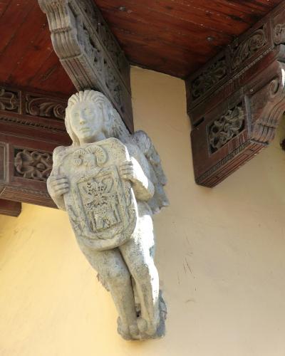 A stone sculpture of a winged figure (putto or angel) holding a coat of arms, mounted under a wooden balcony with carved corbels against a yellow stucco wall.