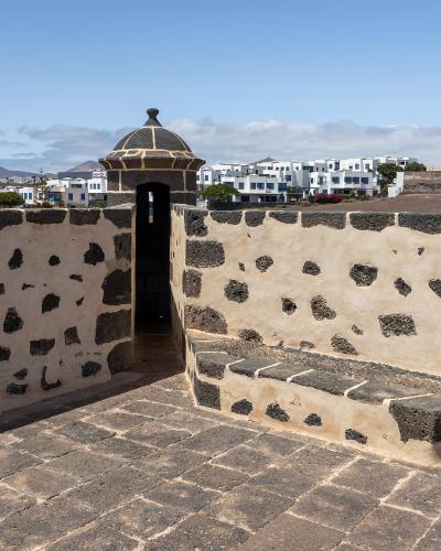 A stone terrace with a distinctive sentry box (garita) featuring a domed roof, overlooking white buildings and volcanic terrain under a blue sky.