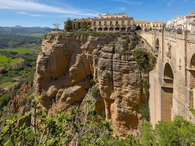 A large stone bridge spanning a deep gorge with a town built on the cliff edge above, featuring a prominent building with arches and a view of green valleys and mountains in the background.