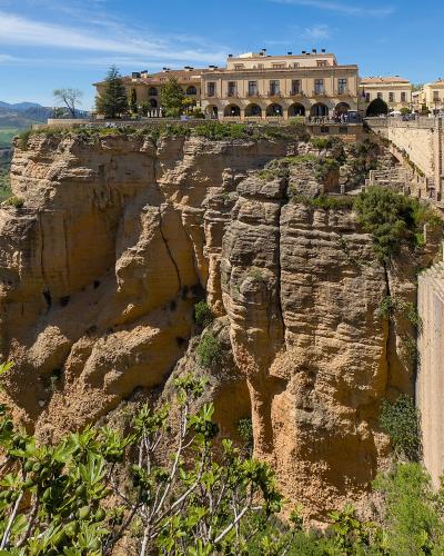 A large stone bridge spanning a deep gorge with a town built on the cliff edge above, featuring a prominent building with arches and a view of green valleys and mountains in the background.