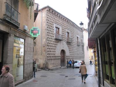 A street scene in a European city showing a distinctive stone building with a facade covered in pyramidal stone protrusions, next to a pharmacy with a green cross sign.