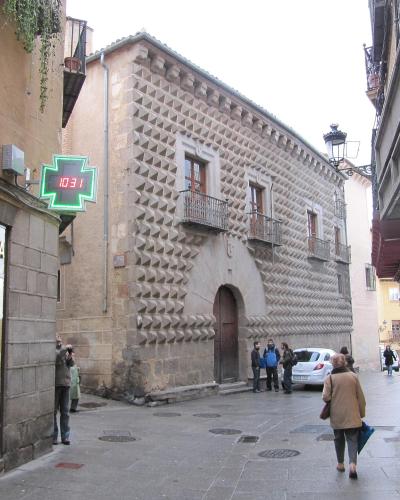 A street scene in a European city showing a distinctive stone building with a facade covered in pyramidal stone protrusions, next to a pharmacy with a green cross sign.