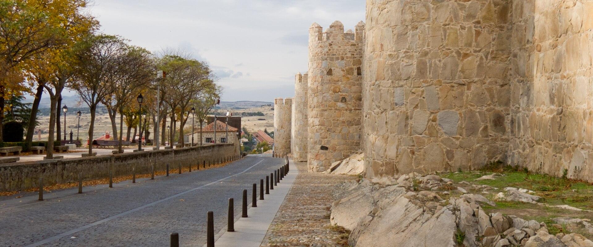 A view of a medieval stone city wall with rounded towers and crenellations, running alongside a paved road and a cobblestone walkway under a partly cloudy sky.