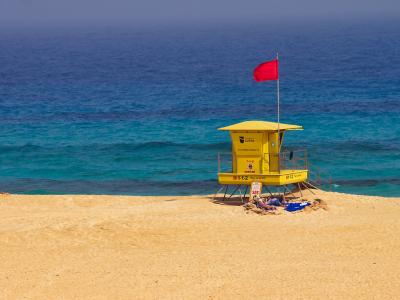 A yellow lifeguard tower stands on a sandy beach with a red flag flying, overlooking turquoise ocean water; text on the tower includes "La Oliva" and emergency numbers.
