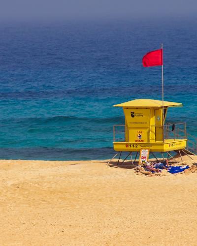 A yellow lifeguard tower stands on a sandy beach with a red flag flying, overlooking turquoise ocean water; text on the tower includes "La Oliva" and emergency numbers.