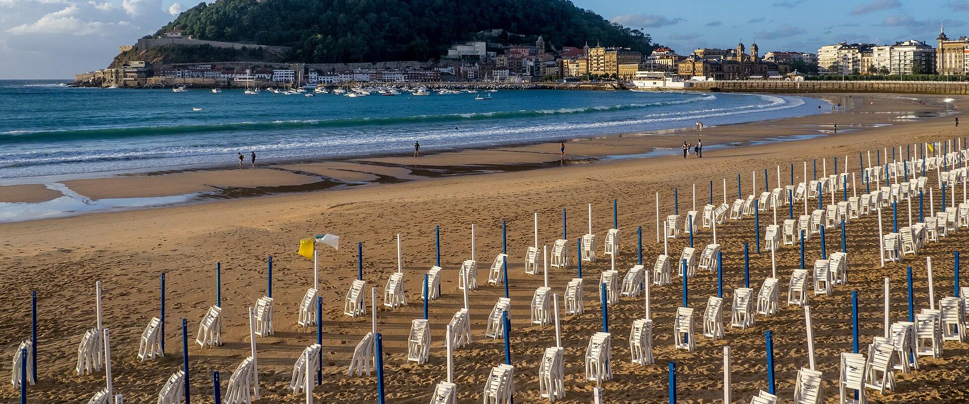 A wide-angle photograph of a sandy beach lined with rows of white wooden deck chairs and blue poles, with a horseshoe-shaped bay, a harbor filled with boats, and a prominent green hill topped with a monument in the background.
