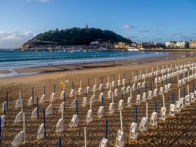 A wide-angle photograph of a sandy beach lined with rows of white wooden deck chairs and blue poles, with a horseshoe-shaped bay, a harbor filled with boats, and a prominent green hill topped with a monument in the background.