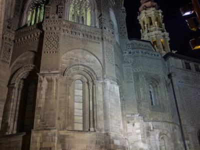 The image shows the illuminated exterior of a large, historic stone cathedral at night, featuring intricate Moorish-style brickwork (mudejar style) on its walls, arched windows, and a prominent bell tower in the background.