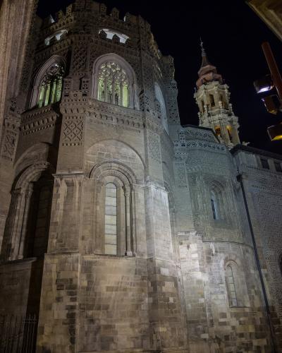 The image shows the illuminated exterior of a large, historic stone cathedral at night, featuring intricate Moorish-style brickwork (mudejar style) on its walls, arched windows, and a prominent bell tower in the background.