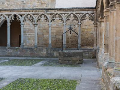 A stone cloister courtyard featuring Gothic-style pointed arches, a central stone well with an iron hand pump, and paved ground with grassy inlays.