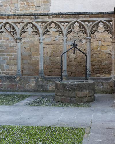 A stone cloister courtyard featuring Gothic-style pointed arches, a central stone well with an iron hand pump, and paved ground with grassy inlays.