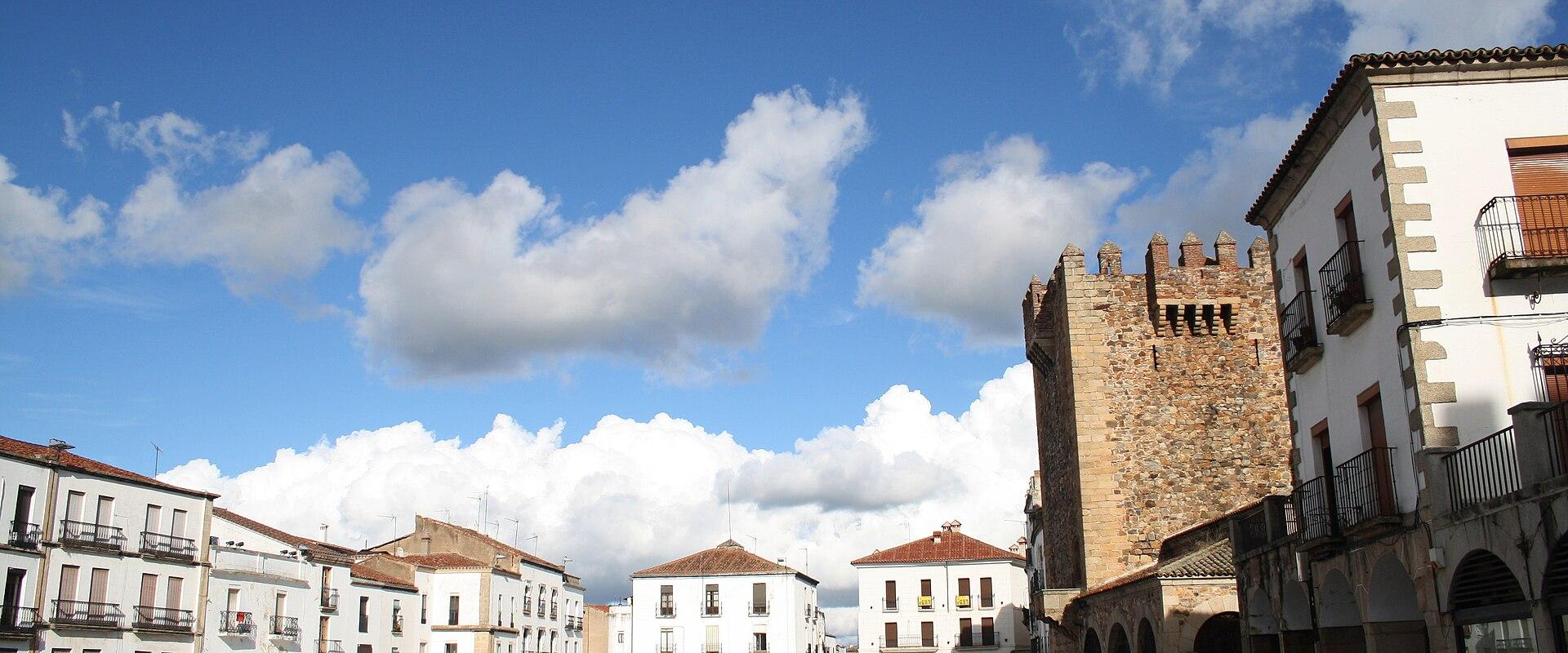 A large, paved public square surrounded by white buildings with wooden balconies and a prominent medieval stone tower with crenellations on the right side.
