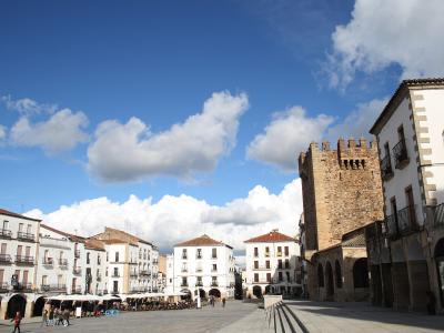 A large, paved public square surrounded by white buildings with wooden balconies and a prominent medieval stone tower with crenellations on the right side.