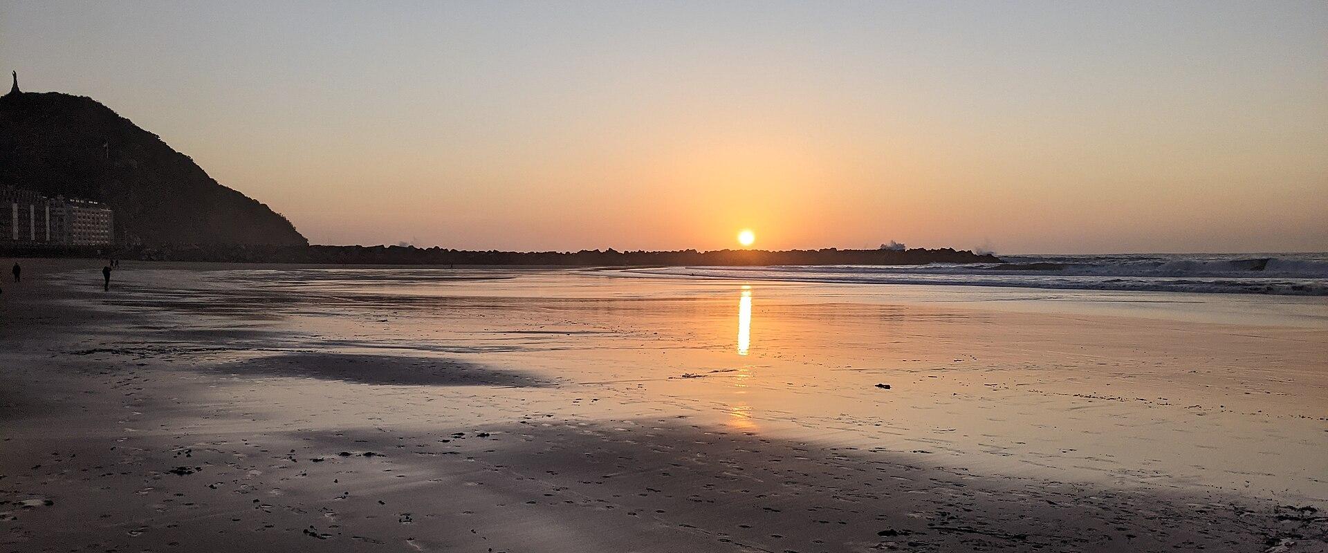 A sunset view of a wide, sandy beach with a large hill (Mount Igueldo) on the left, a line of buildings along the shore, and the sun reflecting on the wet sand.