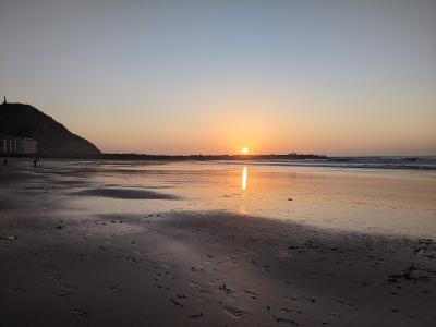 A sunset view of a wide, sandy beach with a large hill (Mount Igueldo) on the left, a line of buildings along the shore, and the sun reflecting on the wet sand.