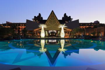 A large swimming pool at twilight reflecting two illuminated, stylized palm tree sculptures and a distinctive building with a central peaked roof and geometric architecture.