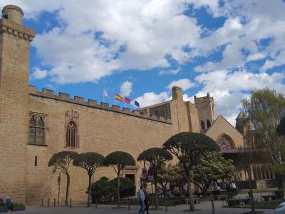 A large, medieval-style stone building with crenellated walls, a prominent square tower, and Gothic-style arched windows, set against a blue sky with clouds, with manicured trees and flags (including the Spanish flag) in front.