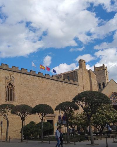A large, medieval-style stone building with crenellated walls, a prominent square tower, and Gothic-style arched windows, set against a blue sky with clouds, with manicured trees and flags (including the Spanish flag) in front.