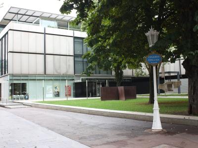 A modern building with white paneling and glass windows, situated next to a park area with trees and a street lamp bearing a blue sign reading "EDUARDO CHILLIDA PLAZA"; text on the building facade reads "Museo de Arte Euskalduna / Museo de Bellas Art