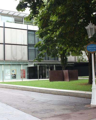 A modern building with white paneling and glass windows, situated next to a park area with trees and a street lamp bearing a blue sign reading "EDUARDO CHILLIDA PLAZA"; text on the building facade reads "Museo de Arte Euskalduna / Museo de Bellas Art