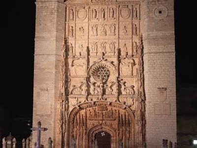 A night photograph of the ornate Gothic stone facade of a large church, featuring two square towers, a central rose window, and a highly detailed sculpted portal, with a stone cross and pillars in the foreground plaza.