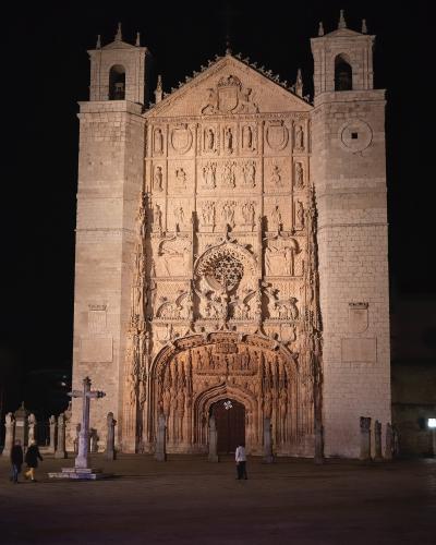 A night photograph of the ornate Gothic stone facade of a large church, featuring two square towers, a central rose window, and a highly detailed sculpted portal, with a stone cross and pillars in the foreground plaza.