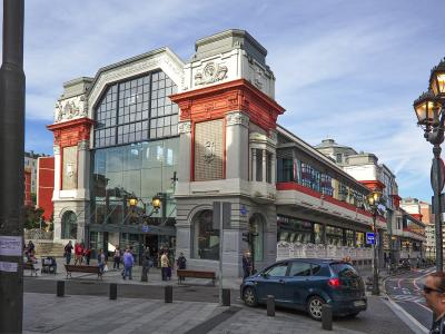 The image shows a large, ornate market building with distinctive red and grey architecture, large glass windows, and people walking on the sidewalk in front of it.