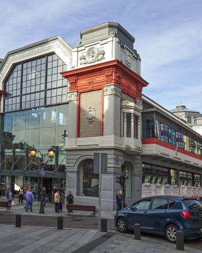The image shows a large, ornate market building with distinctive red and grey architecture, large glass windows, and people walking on the sidewalk in front of it.