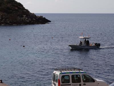 A white utility van with green and blue stripes is parked in the foreground overlooking a bay with a rocky headland on the left and a small motorboat moving across the water.