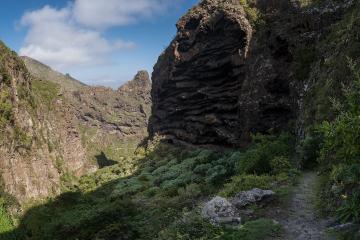 A dirt hiking trail winds along the side of a steep, rocky ravine with layered volcanic rock formations and lush green vegetation under a blue sky.