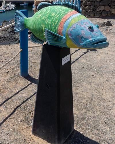 A large, colorful sculpture of a fish mounted on a black pedestal, with a harbor and stone wall in the background.