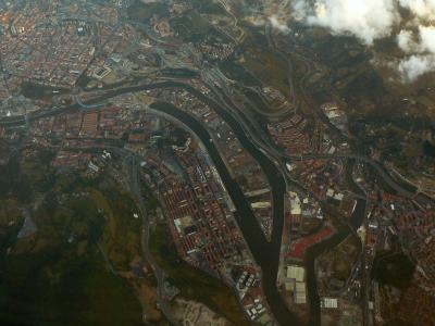 An aerial photograph showing a wide river estuary flowing through a dense urban area with extensive road networks, bridges, and industrial zones, under cloudy skies.