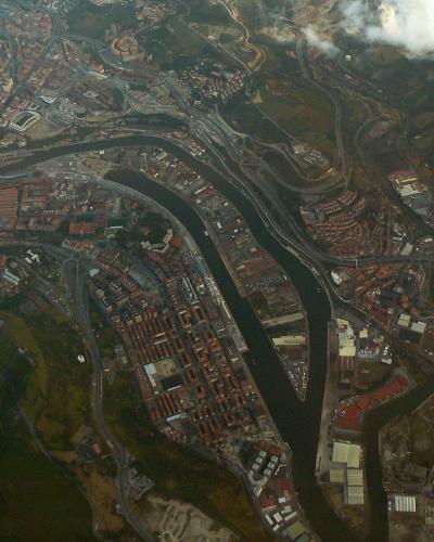 An aerial photograph showing a wide river estuary flowing through a dense urban area with extensive road networks, bridges, and industrial zones, under cloudy skies.