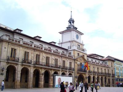 The image shows a large, historic stone building with a prominent clock tower, arched ground-floor colonnades, and balconies, situated in a public square with people walking around.