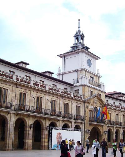 The image shows a large, historic stone building with a prominent clock tower, arched ground-floor colonnades, and balconies, situated in a public square with people walking around.