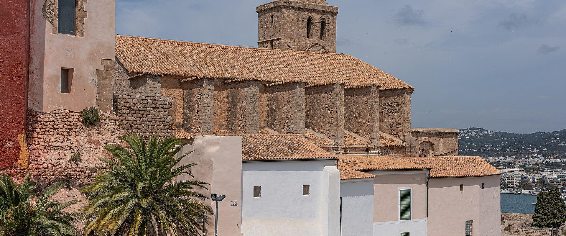 A stone church with a distinctive octagonal bell tower and terracotta tiled roof, situated on a hillside above white buildings and palm trees, with a view of a coastal town and harbor in the background.