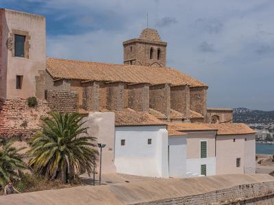 A stone church with a distinctive octagonal bell tower and terracotta tiled roof, situated on a hillside above white buildings and palm trees, with a view of a coastal town and harbor in the background.