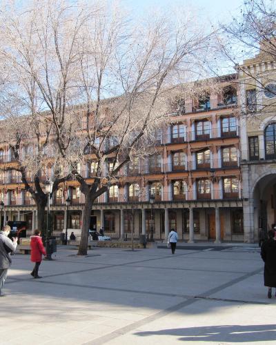 A wide, paved public square lined with multi-story buildings featuring arcades and balconies, with bare trees and pedestrians.