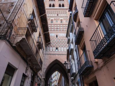 A low-angle view of a narrow cobblestone street in a historic Spanish town, looking up at a tall, ornate brick tower with Mudéjar-style geometric tilework and an archway at its base.