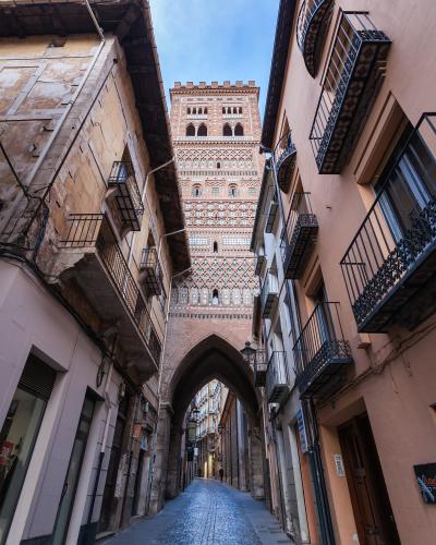 A low-angle view of a narrow cobblestone street in a historic Spanish town, looking up at a tall, ornate brick tower with Mudéjar-style geometric tilework and an archway at its base.