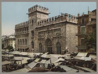 A historic stone building with Gothic architecture, crenellated towers, and arched windows, fronted by a bustling outdoor market with numerous white awnings.