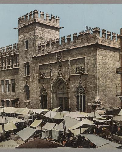 A historic stone building with Gothic architecture, crenellated towers, and arched windows, fronted by a bustling outdoor market with numerous white awnings.