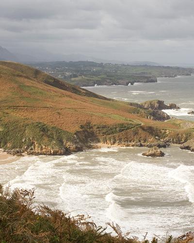A coastal landscape showing a sandy beach nestled at the base of a grassy, sloping hill with a winding path, rocky outcrops in the surf, and distant mountains under an overcast sky.