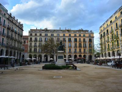 A wide, open public square with a central equestrian statue on a stone pedestal, surrounded by multi-story buildings with arched ground-floor arcades and balconies, under a cloudy sky.