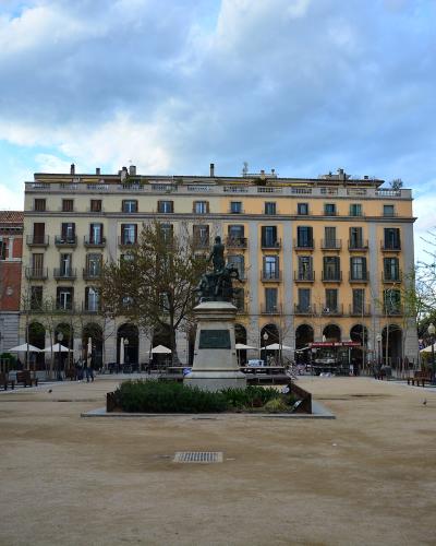 A wide, open public square with a central equestrian statue on a stone pedestal, surrounded by multi-story buildings with arched ground-floor arcades and balconies, under a cloudy sky.