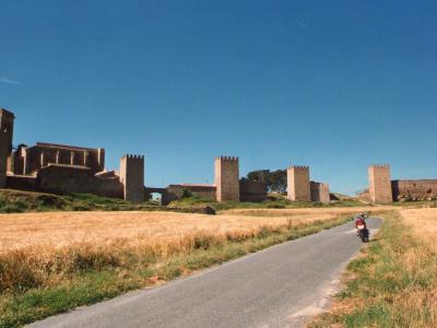 A medieval stone fortress with crenellated towers and walls, situated on a grassy hill overlooking a paved road and golden fields under a clear blue sky.
