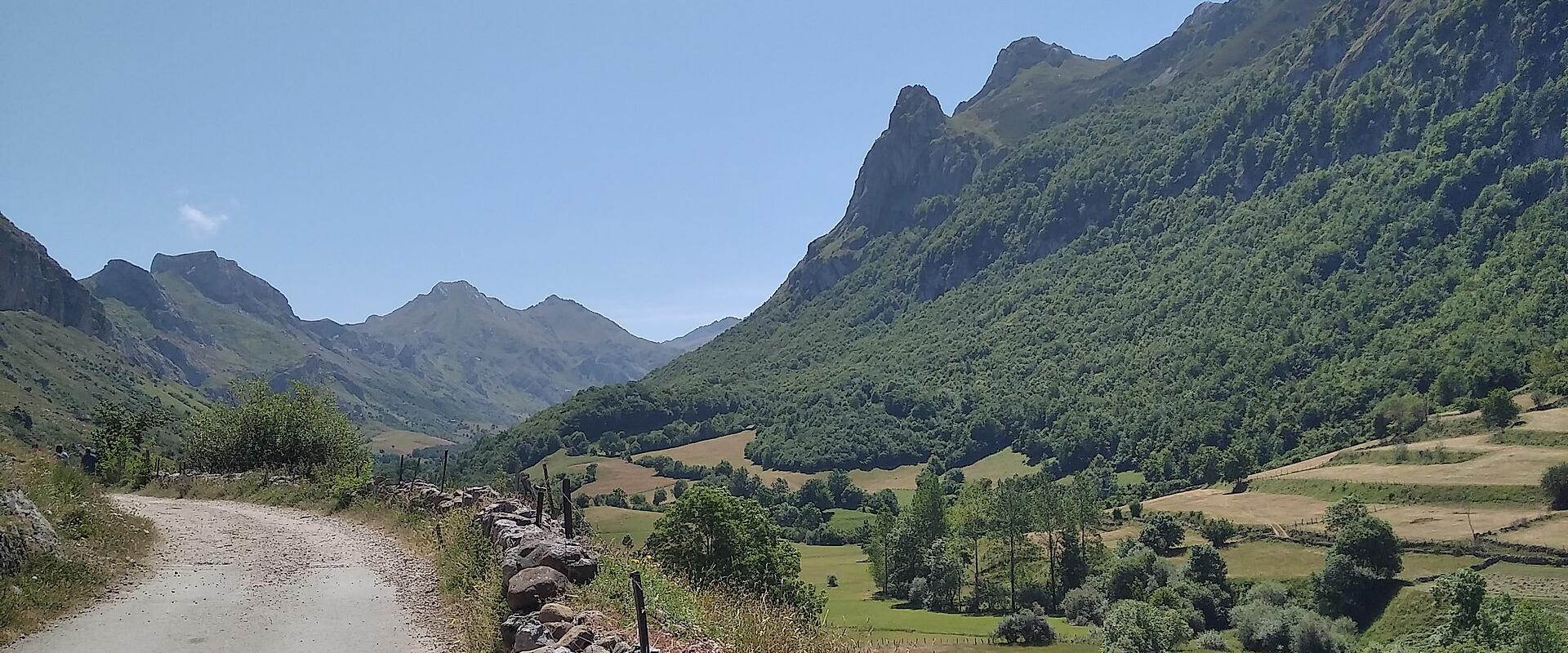 A dirt road runs alongside a dry-stone wall through a green, mountainous valley with rolling hills and forested slopes under a clear blue sky.