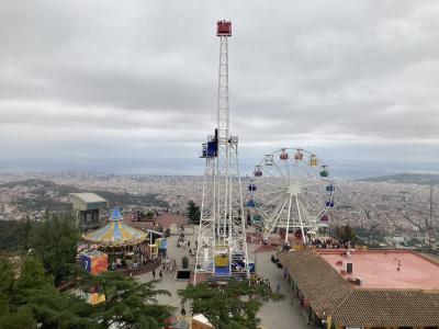 An amusement park featuring a tall white drop tower ride, a large Ferris wheel, and a carousel, set on a hilltop overlooking a dense urban cityscape and the sea.